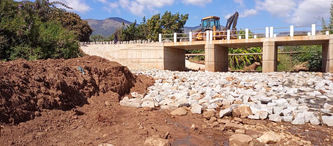 Ponte concluída com retroescavadora no tabuleiro e aterro de acesso em terra vermelha